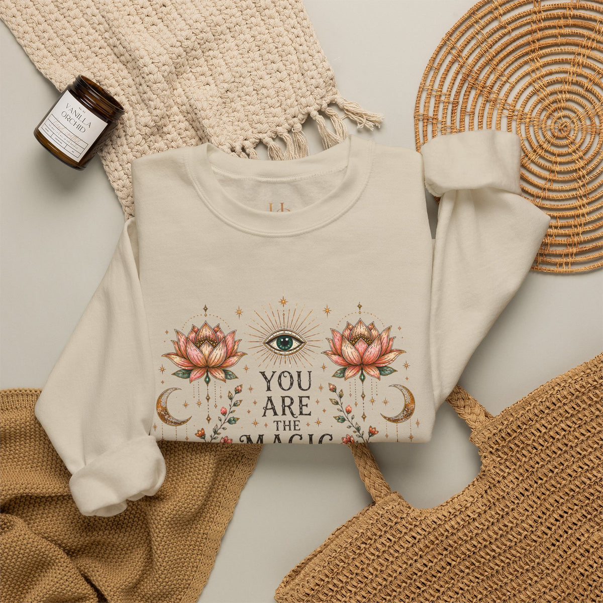 An off-white long-sleeve crewneck sweatshirt with a bohemian-style graphic print featuring a green eye, pink lotus flowers, gold crescent moons, and the text 'YOU ARE THE MAGIC'. The sweatshirt is styled in a flat-lay arrangement with a cream knit blanket, a brown candle jar, and a woven straw mat on a light gray background.