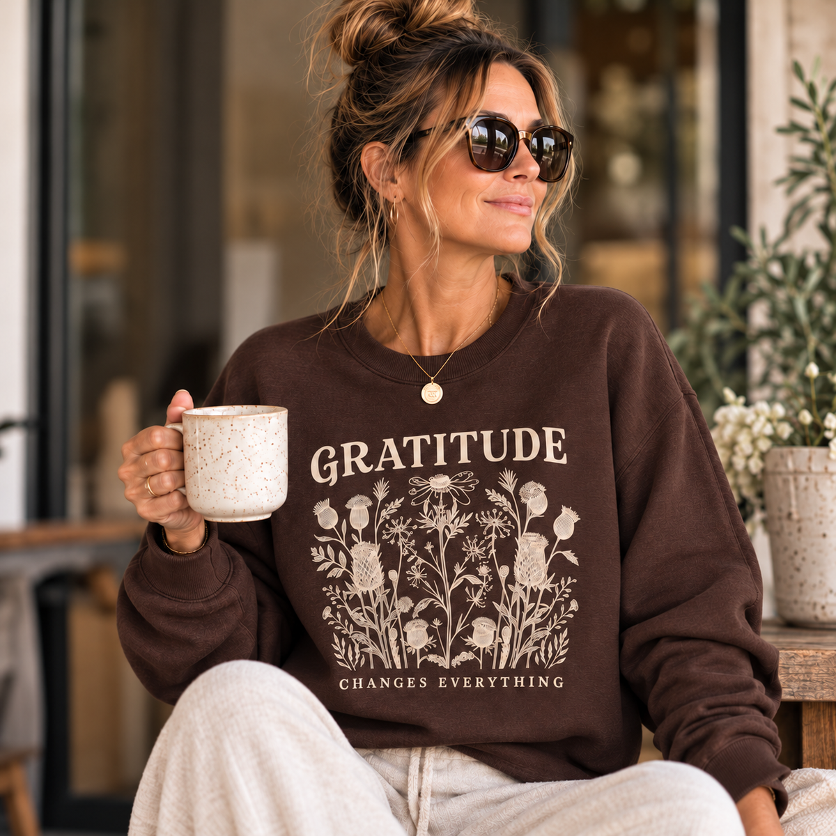 Woman wearing the dark chocolate Gratitude Changes Everything floral sweatshirt with cream joggers, holding a coffee mug