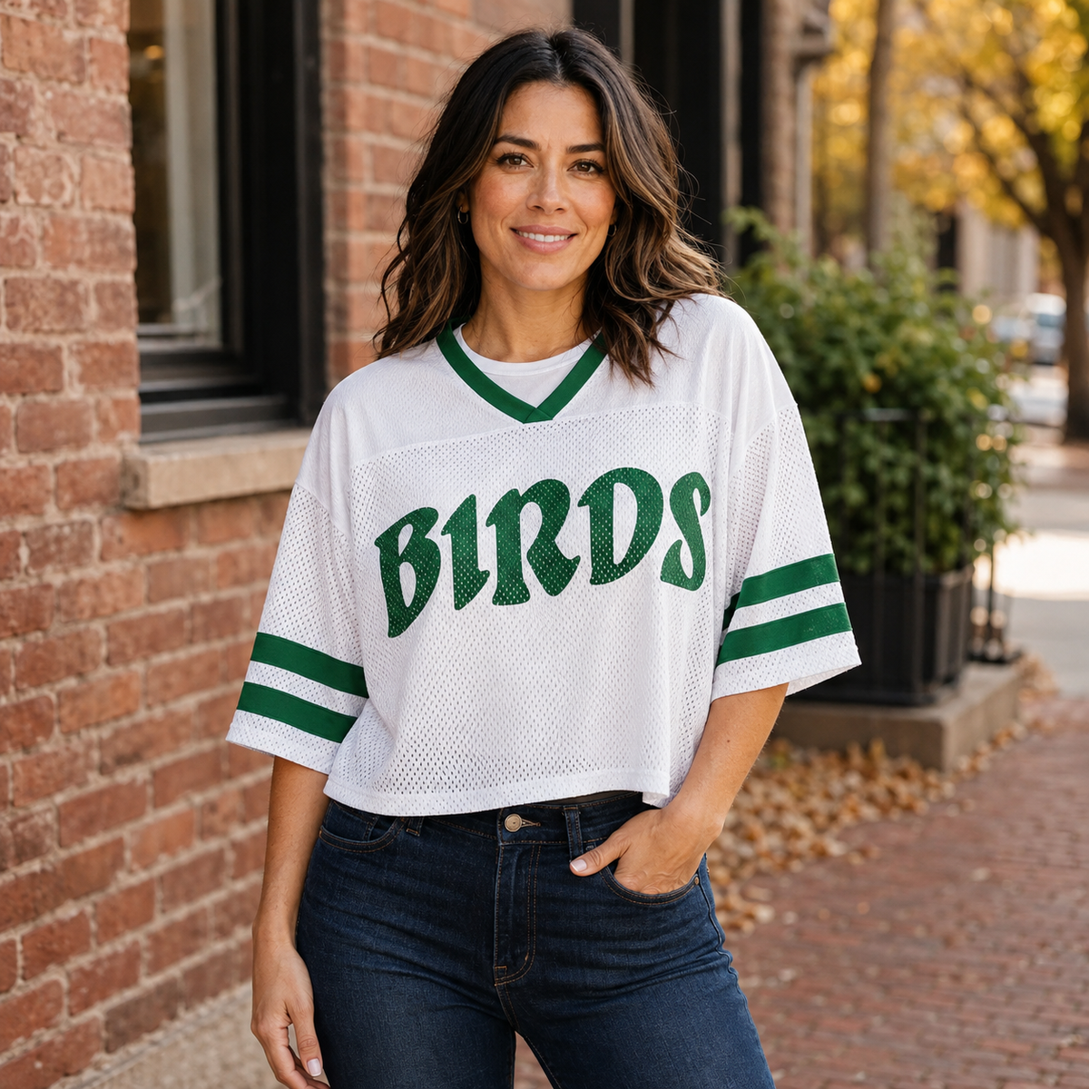 Woman wearing a white cropped mesh B1RDS jersey with Kelly green V-neck and sleeve stripes on a Philadelphia brick street.